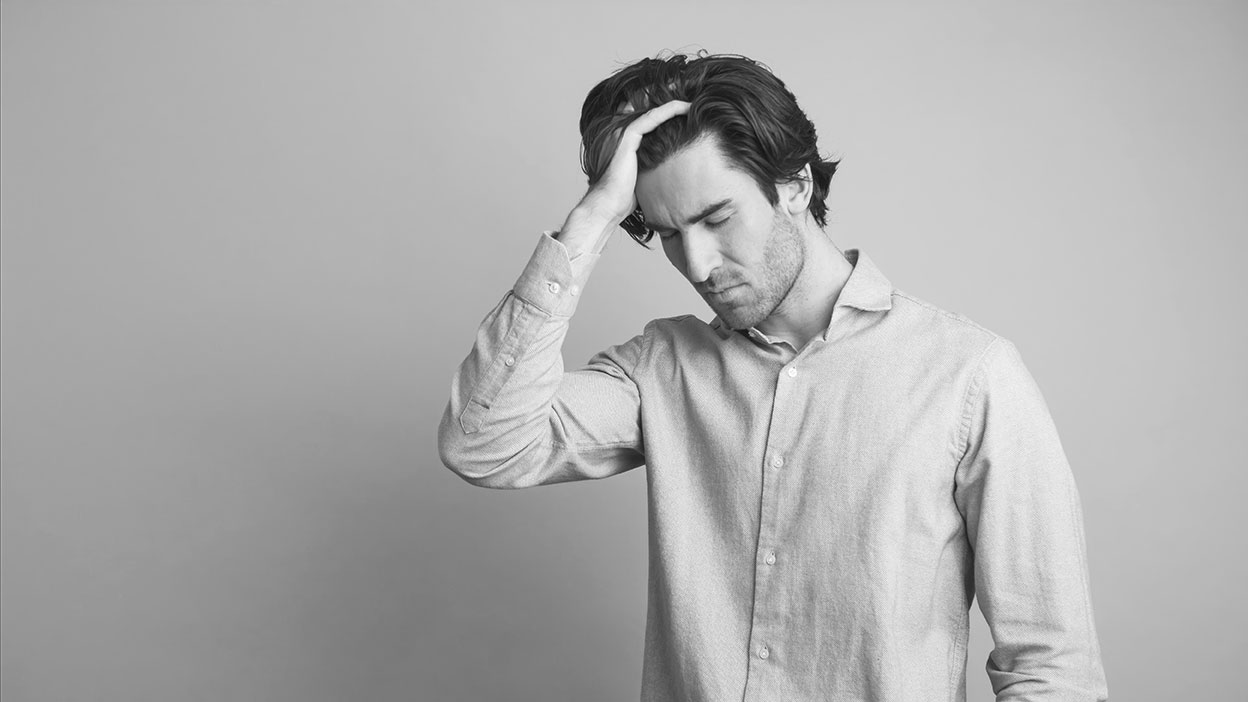 nuka-ai-app-overload-stressed-man Schwarz-weiß-Foto eines jungen Mannes mit längeren dunklen Haaren, der ein Hemd trägt. Er steht vor einem neutralen Hintergrund und wirkt innerlich angespannt oder überfordert. Mit der rechten Hand fährt er sich durch die Haare und hat die Augen gesenkt. Seine Körperhaltung und Mimik drücken Nachdenklichkeit, Erschöpfung oder mentale Belastung aus.